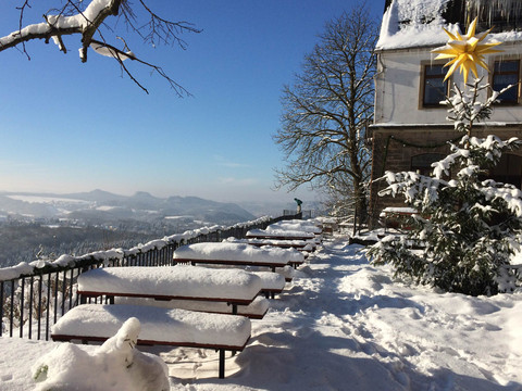 Verschneite Terrasse mit Holzbänken und Tischen, ein geschmückter Tannenbaum mit gelbem Stern, blauer Himmel und schneebedeckte Landschaft im Hintergrund.