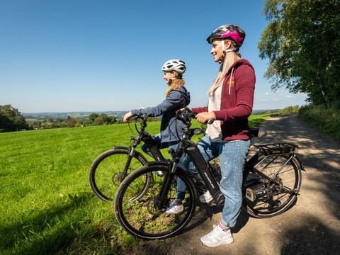Heckberg Zwei Radfahrer mit Helmen genießen die Aussicht auf eine grüne, hügelige Landschaft bei Sonnenschein.