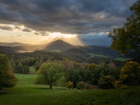 Blick zum Rosenberg Berglandschaft bei Sonnenuntergang mit dramatischen Wolken, Sonnenstrahlen, grünen Wiesen und bunten Bäumen im Vordergrund.Mountain landscape at sunset with dramatic clouds, sunbeams, green meadows and colorful trees in the foreground.Horská krajina při západu slunce s dramatickými mraky, slunečními paprsky, zelenými loukami a barevnými stromy v popředí.Górski krajobraz o zachodzie słońca z dramatycznymi chmurami, promieniami słońca, zielonymi łąkami i kolorowymi drzewami na pierwszym planie.Berglandschap bij zonsondergang met dramatische wolken, zonnestralen, groene weiden en kleurrijke bomen op de voorgrond.Paesaggio montano al tramonto con nuvole, raggi di sole, prati verdi e alberi colorati in primo piano.