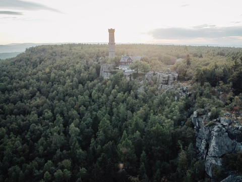 Ein hoher Turm und ein Gebäude stehen auf einem bewaldeten Hügel, umgeben von dichtem Grün und Felsen, bei sanftem Sonnenlicht.