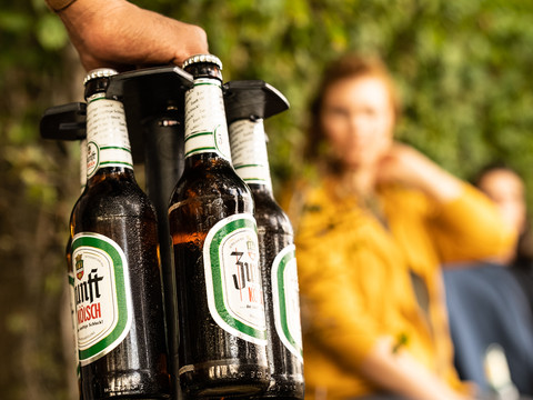 Bierweg Bierflaschen auf einem Flaschenhalter im Vordergrund, unscharfe Personen im Hintergrund.