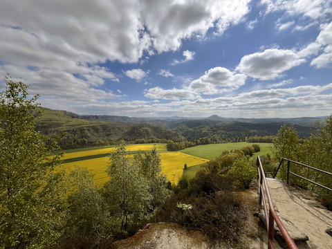 Aussicht auf eine weite Landschaft mit gelben Rapsfeldern, grünen Wiesen und Hügeln unter einem bewölkten blauen Himmel, im Vordergrund ein Geländer.