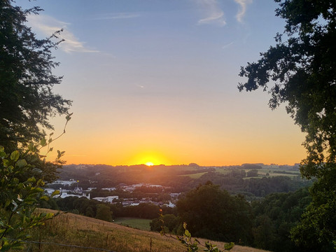 Overather Panoramaweg Sonnenuntergang über einer hügeligen Landschaft, umgeben von Bäumen und Feldern, Himmel orange leuchtend.