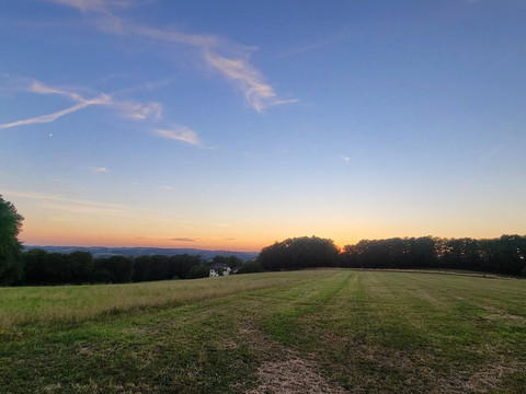 Overather Panoramaweg Sonnenuntergang über einer ruhigen, ländlichen Wiese mit Feldern und Bäumen im Hintergrund.
