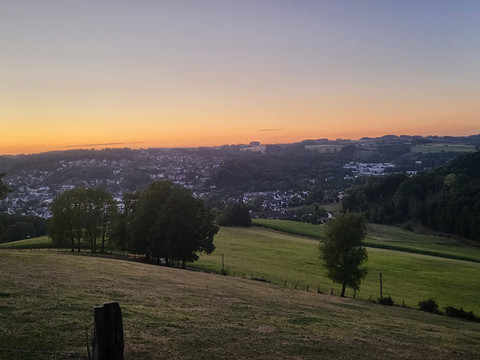 Overather Panoramaweg Panoramablick auf eine sanft hügelige Landschaft bei Sonnenuntergang, mit kleinen Städten im Hintergrund.
