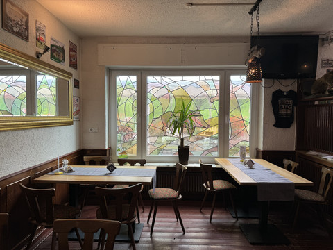 Zur Quelle der Strunde - Innenansicht Gemütliches Café mit Holzstühlen, buntem Fenster und Pflanzen auf einem Tisch. Wanddeko rundet das Bild ab.