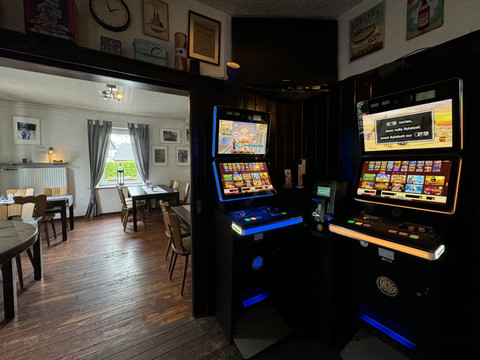 Zur Quelle der Strunde - Spielautomaten <p>Spielautomaten in einer gemütlichen Ecke eines rustikalen Gastraums mit Holzboden und Tischgarnituren.</p>