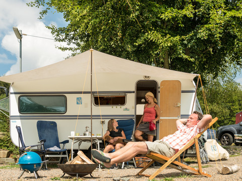 Campingpark im Bergischen Land Ein Paar entspannt vor einem Wohnwagen, Frau steht in Tür. Campingplatz bei sonnigem Wetter und Bäumen.