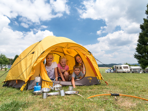 Campingpark im Bergischen Land Zelt auf Wiese mit Kindern, Campingplatz und Wohnwagen im Hintergrund, sonniger Himmel.