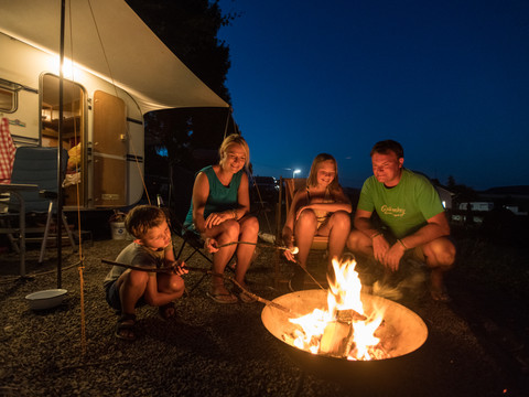 Campingpark im Bergischen Land Ein Camper-Familienlagerfeuer bei Nacht, Menschen braten Marshmallows, im Hintergrund ein Wohnwagen.