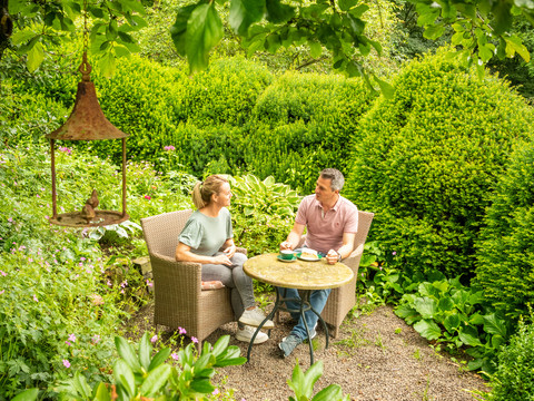 Ferienwohnung Hof Tüschenbonnen Zwei Personen sitzen im grünen Garten an einem runden Tisch und genießen eine Tasse Kaffee.