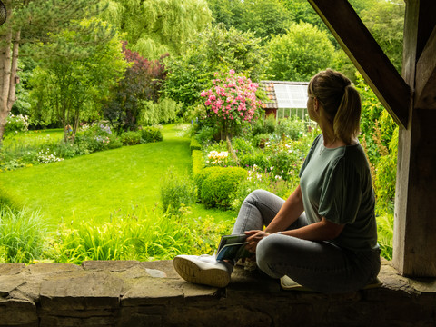 Ferienwohnung Hof Tüschenbonnen <p>Frau auf Steinmauer blickt in blühenden Garten mit Gewächshaus im Hintergrund.</p>
