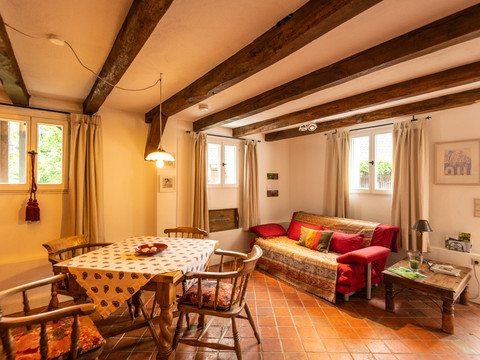 Ferienwohnung Hof Tüschenbonnen Gemütliches Wohnzimmer mit Holzbalkendecke, Esstisch, rotem Sofa, Fenster mit Vorhängen und Fliesenboden.