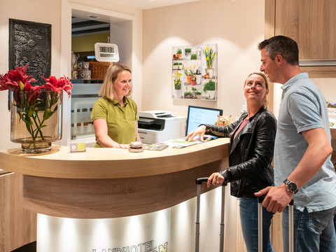 Landhaus Napoleon An der Rezeption des Landhotels begrüßt eine Mitarbeiterin freundlich ein lächelndes Paar mit Koffern.