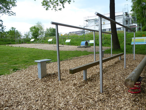 Fitnessparcours Barrierefreier Fitnesspark mit Holzschnitzelboden, Balancierbalken, Reckstangen und Begrünung im Hintergrund.
