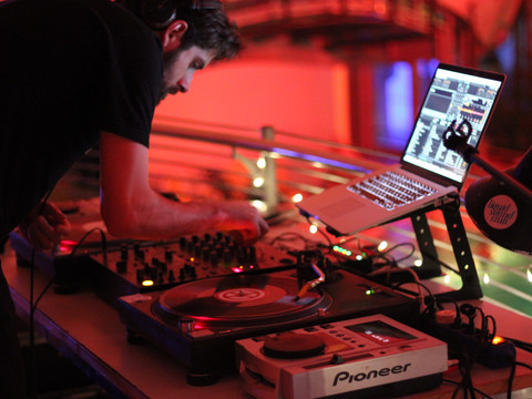 Liquid Sound Club Ein DJ performt in einem gedämpft beleuchteten Raum mit moderner Ausrüstung, umgeben von rotem Licht.A DJ performs in a dimly lit room with modern equipment, surrounded by red light.V tlumeně osvětlené místnosti s moderním vybavením, obklopené červeným světlem, hraje DJ.DJ występuje w słabo oświetlonym pomieszczeniu z nowoczesnym sprzętem, otoczony czerwonym światłem.Een DJ treedt op in een schemerige kamer met moderne apparatuur, omgeven door rood licht.Un DJ si esibisce in una stanza poco illuminata con attrezzature moderne, circondato da luci rosse.