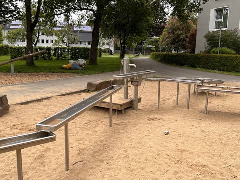 Wasserspielplatz im Kurpark Barrierefreier Spielplatz mit Sand und Edelstahl-Wasserspiel in einer grünen Parkanlage vor Gebäuden.