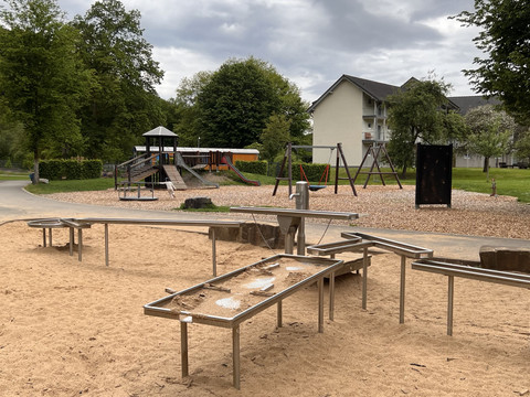 Wasserspielplatz im Kurpark Spielplatz mit Sandbereich, Holzrutschen und Schaukeln, umgeben von Bäumen und einem weißen Gebäude.