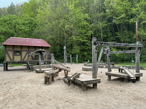 Café Alte Dombach - Ansicht Spielplatz Ein rustikaler Spielplatz mit Holzhäuschen und Spielgeräten, umgeben von üppigem Wald.