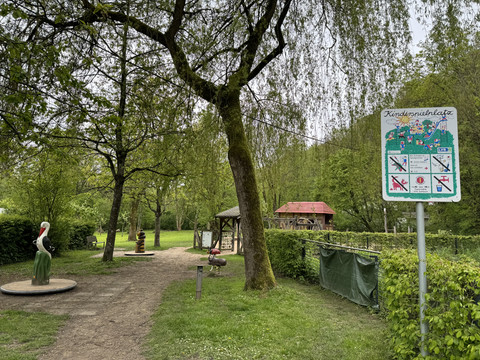 Café Alte Dombach - Ansicht Spielplatz <p>Spielplatz im Grünen mit Holzskulpturen, Spielgerät und Hinweisschild, eingebettet in Bäume und Sträucher.</p>