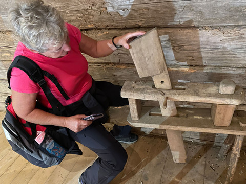 Eine Frau in einem pinken T-Shirt betrachtet ein altes hölzernes Werkzeug in einer rustikalen Holzhütte.