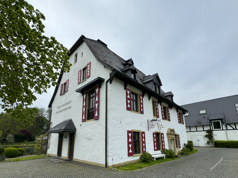 Außenansicht Fachwerkhaus mit weißen Wänden, roten Fensterläden und Schieferdach, umgeben von gepflastertem Hof.