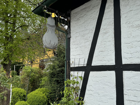 Schild der Dröppelminna Fachwerkhaus mit weißer Fassade und schwarzen Balken. Daneben gepflegter Garten mit grünem Laub.