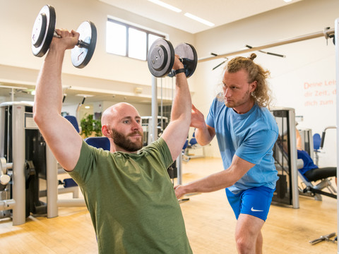 Trans World Hotel Kranichhöhe <p>Ein Mann hebt Hanteln im Fitnessstudio, während ein Trainer ihm hilft.</p>