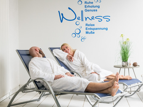 Das Bergische-2020-100-Gesundheit.jpg Entspannte Menschen in Bademänteln liegen auf Wellnessliegen im Spa-Bereich und lächeln einander an.