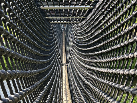 Seilbrücke auf dem Abenteuerspielplatz Ein schmaler, schwankender Hängebrückenkorridor aus Seilen, der über einen grünen Park führt.