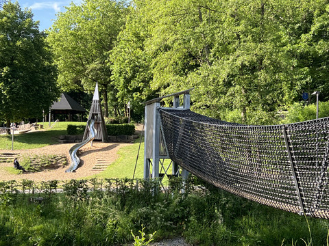 Abenteuerspielplatz im Wiehlpark Eine Spielplatzlandschaft mit einer Rutsche aus Holz und einer Hängebrücke, umgeben von grünen Bäumen.