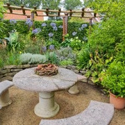 Sitzecke Garten Angelika und Holger Ihler.jpg Das Bild zeigt eine idyllische Gartensitzecke, die von üppigem Grün und blühenden Pflanzen umgeben ist. Ein runder Steintisch mit zwei halbmondförmigen Steinbänken wird von einem rustikalen Gartenzaun eingerahmt. Die farbenfrohe Blumenvielfalt und das satte Grün der Blätter schaffen eine friedliche und einladende Atmosphäre.