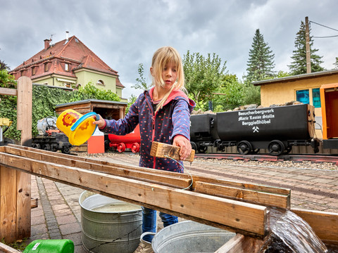 Am Besucherbergwerk Kind spielt mit Wasser am Besucherbergwerk vor historischen Loren und grüner Landschaft.