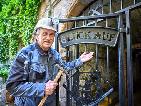 Glück Auf! Am Besucherbergwerk "Marie Louise Stolln" Ein lächelnder Bergmann in Arbeitskleidung zeigt auf ein Grubentor mit dem Schriftzug "Glück Auf!".