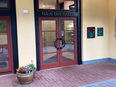 Haus des Gastes Eingang Eingang des "Haus des Gastes" mit roten Türen, Blumentopf mit bunten Blumen links, Infotafeln an der Wand.Entrance to the "Haus des Gastes" with red doors, flower pot with colorful flowers on the left, information boards on the wall.Vchod do "Haus des Gastes" s červenými dveřmi, vlevo květináč s barevnými květinami, na stěně informační tabule.Wejście do "Haus des Gastes" z czerwonymi drzwiami, donica z kolorowymi kwiatami po lewej stronie, tablice informacyjne na ścianie.Ingang van het "Haus des Gastes" met rode deuren, bloempot met kleurrijke bloemen aan de linkerkant, informatieborden aan de muur.Ingresso della "Haus des Gastes" con porte rosse, vaso di fiori colorati sulla sinistra, pannelli informativi sulla parete.