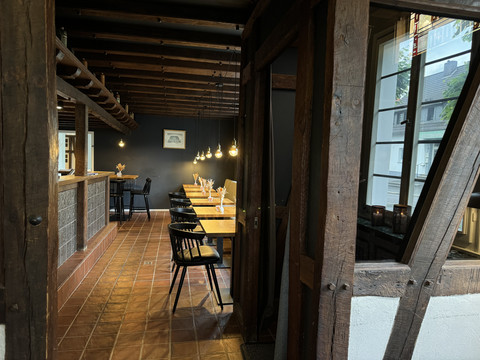Zum Haus Olpe - Ansicht Innenbereich  Gemütliches Restaurant mit Holzbalken, langen Holztischen und warmem Licht von hängenden Lampen.
