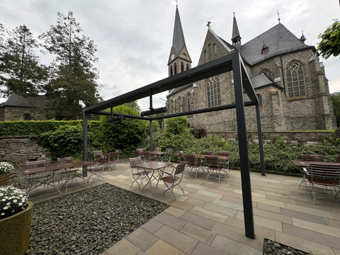 Zum Haus Olpe - Ansicht Außenbereich Kirche mit zwei Türmen, blühendem Garten und Sitzbereich aus Holz vor bewölktem Himmel in ruhiger Umgebung.