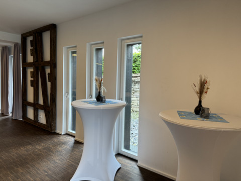Zum Haus Olpe - Innenansicht Veranstaltungsraum  Heller Raum mit Stehtischen und schlichten Dekorationen, modernen Fenstern und einem rustikalen Holzelement.