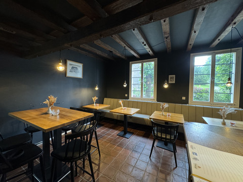 Zum Haus Olpe - Ansicht Innenbereich  Gemütlicher Innenraum eines Cafés mit Holztischen, schwarzen Stühlen und weichen Bänken, beleuchtet durch warmes Licht.