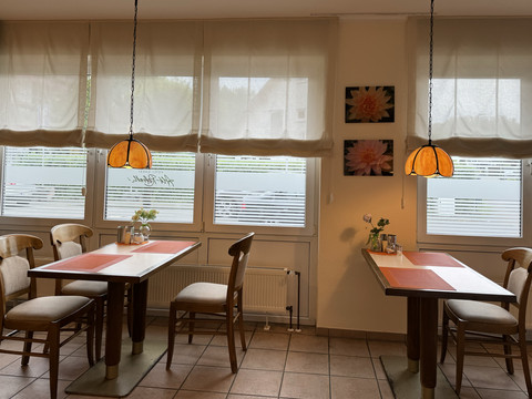 Hotel Garni Alt Refrath - Innenansicht Essbereich  Gemütliches Café mit Holztischen, Stühlen und warmen Lampen vor großen, lichtdurchfluteten Fenstern.