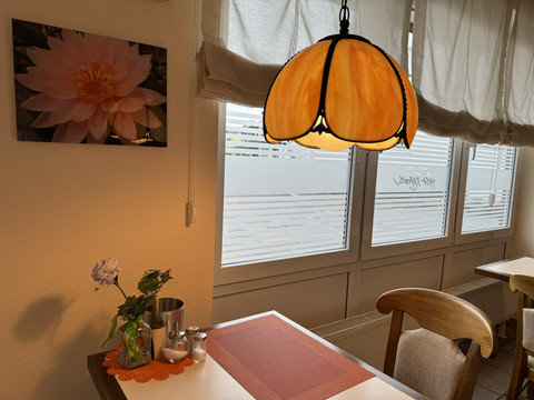 Hotel Garni Alt Refrath - Innenansicht Essbereich Eine gemütliche Café-Ecke mit orangener Lampe, Blume und Tischset, von Tageslicht beleuchtet.