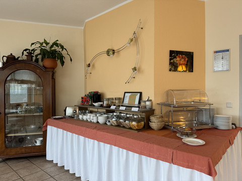 Hotel Garni Alt Refrath - Ansicht Essbereich <p>Frühstücksbuffet mit Müsli, Brotkorb und Geschirr, neben einem antiken, verglasten Holzschrank.</p>