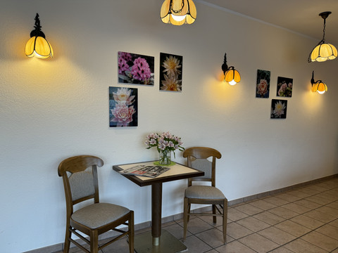 Hotel Garni Alt Refrath - Ansicht Essbereich  <p>Zwei Holzstühle an einem Tisch, Blumen und Bilder an weißer Wand; gemütliche Atmosphäre.</p>