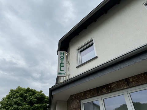 Hotel Garni Alt Refrath - Außenansicht Hotelgebäude mit grünem "Hotel Garni"-Schild an der Fassade, bewölkter Himmel im Hintergrund.