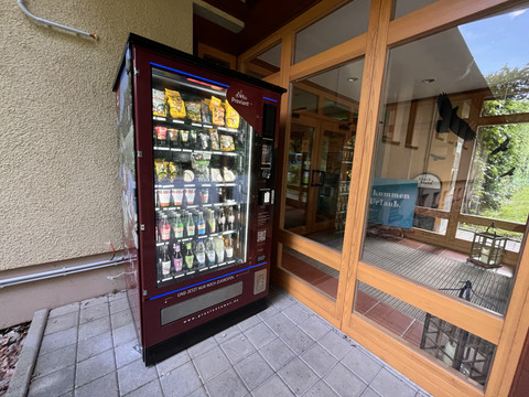 Ein roter Verkaufsautomat mit Snacks und Getränken steht neben einer Glastür; im Hintergrund ist ein Schild mit "Willkommen im Urlaub" zu sehen.
