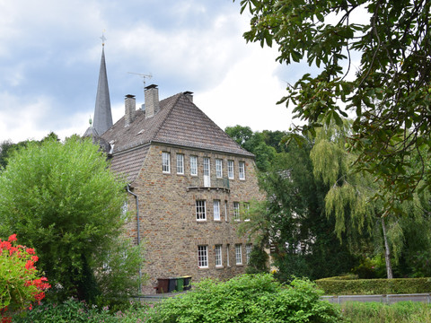 Ensemble Volberg  Altes Steinhaus mit mehreren Schornsteinen und einem Kirchturm im Hintergrund, umgeben von grüner Vegetation.