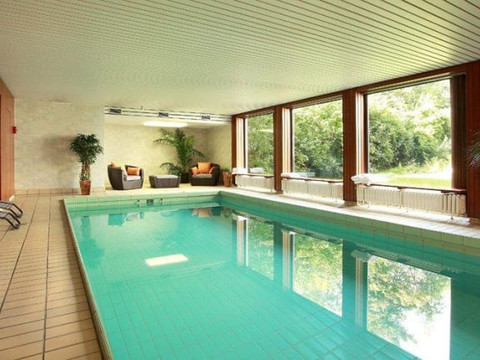 Schwimmbad.jpg Innenansicht eines modernen Hallenbades mit großen Fenstern, Topfpflanzen und gemütlichen Sitzmöbeln.