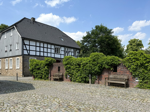 IMG_1109.jpeg Fachwerkhaus mit grauem Dach, umgeben von Bäumen, Backsteinmauer und zwei Holzbänken auf Pflastersteinen.