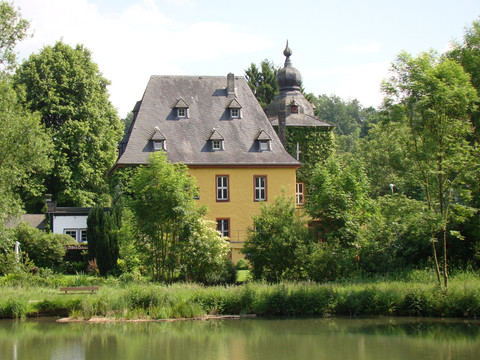 Bergischer Weg Etappe 6 Gelbes Haus mit Schieferdach am Seeufer, umgeben von üppigem Grün, im Sommer unter blauem Himmel.