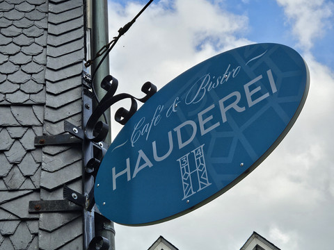 Hauderei Café & Bistro Das Bild zeigt ein blaues, ovales Schild mit der Aufschrift "Café & Bistro Hauderei" vor einem Haus.
